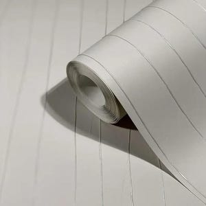 May include: A roll of white wallpaper with vertical lines and thin silver stripes. The wallpaper is partially unrolled, revealing the rolled-up section. The design features a subtle texture and a modern aesthetic, suitable for interior decoration.