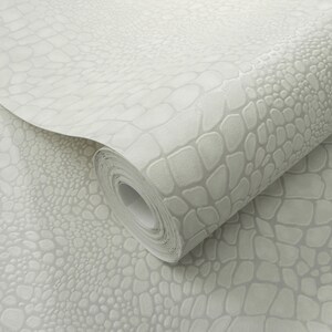 May include: A roll of light gray wallpaper with a textured, embossed pattern resembling crocodile or alligator skin. The wallpaper is partially unrolled, revealing the pattern and the paper's rolled form. The design is a neutral color, suitable for various interior styles.