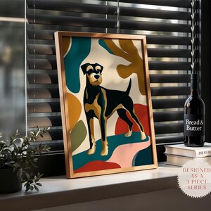Puede incluir: Una impresión artística enmarcada con un perro negro y fuego estilizado sobre un fondo de formas abstractas en turquesa, dorado y rojo. La impresión está en un marco dorado, exhibida en un alféizar con una botella de vino y libros.