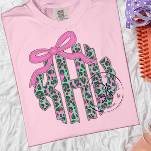 May include: A light pink t-shirt featuring a pink bow and the letters "RHB" in a green and pink leopard print design. The shirt is made of a soft material and is perfect for casual wear.