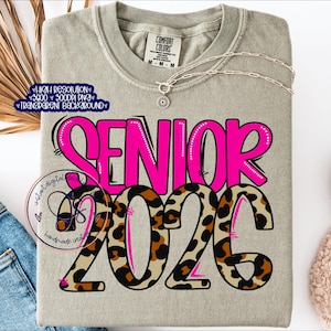 May include: A light gray Comfort Colors t-shirt with the word "SENIOR" in bright pink, outlined in black. The numbers "2025" are in a leopard print design. A silver necklace with a pendant is draped across the shirt.