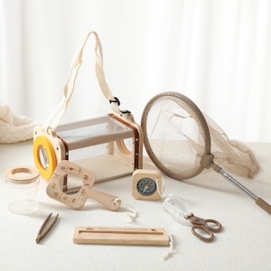 May include: A collection of wooden outdoor exploration toys, including a bug catcher with a handle, a magnifying glass, a compass, a net, tweezers, and scissors. The toys are made of natural wood and light-colored materials.