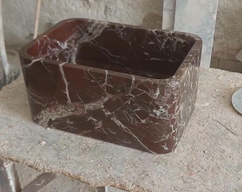 May include: A rectangular, dark red marble sink with white veining. The sink has rounded corners and a smooth, polished surface. The bathroom fixture is set on a light-colored, textured surface.