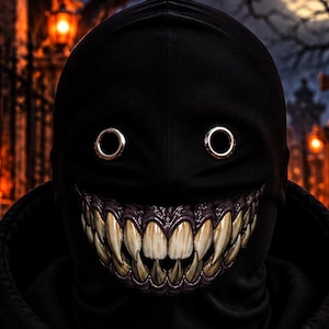 Op de afbeelding: Een zwarte hoodie met een masker met twee ronde oogopeningen en een brede, tandige grijns. Het masker toont scherpe, vergeelde tanden en donker tandvlees. Het woord OTKYDACH is in witte, druipende letters op de borst van de hoodie gedrukt.