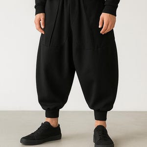 May include: A person in an all-black outfit: a crew neck sweatshirt, wide-leg trousers with pockets, and trainers. They are wearing a black balaclava. The ensemble is minimalist and contemporary.