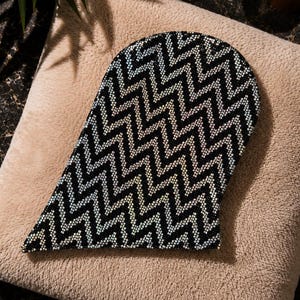 May include: A curved exfoliating mitt featuring a dense, repeating black and white chevron pattern. The surface is covered in small, textured beads that create a distinct zigzag design across the entire front of the mitt, providing a tactile finish.