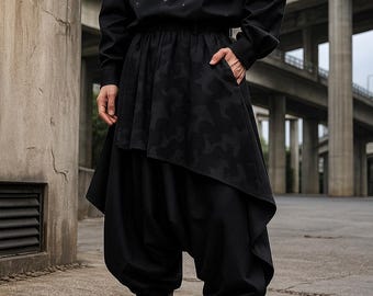 May include: A person in a black outfit, featuring a long-sleeved shirt with white paint splatters, a layered skirt with a camouflage pattern, and baggy trousers. The outfit is completed with black boots.