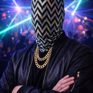 May include: A person wearing a black jacket, a black shirt, and a gold chain necklace. The person is wearing a mask with a black and silver chevron pattern. The background is a nightclub with colorful lights. neo robot