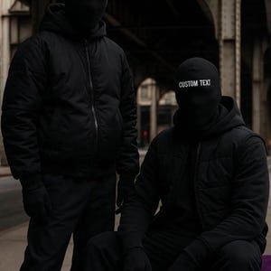 May include: Two people wearing black balaclavas with "CUSTOM TEXT" printed in white. They are dressed in black jackets, pants, and gloves. One person is standing, and the other is seated on a purple object, all against an urban backdrop. neo robot