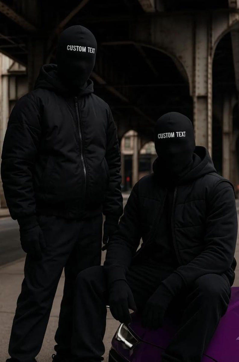 May include: Two people wearing black balaclavas with "CUSTOM TEXT" printed in white. They are dressed in black jackets, pants, and gloves. One person is standing, and the other is seated on a purple object, all against an urban backdrop. neo robot