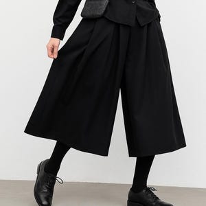 May include: A person wearing a black outfit, including a button-down shirt, wide-leg pants, and leather shoes. A small black crossbody bag is also visible. The outfit is all black.
