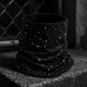 May include: A black, cylindrical neck warmer made of soft fabric, adorned with numerous sparkling rhinestones. The neck warmer has a ruched design and is displayed on a textured, dark surface.