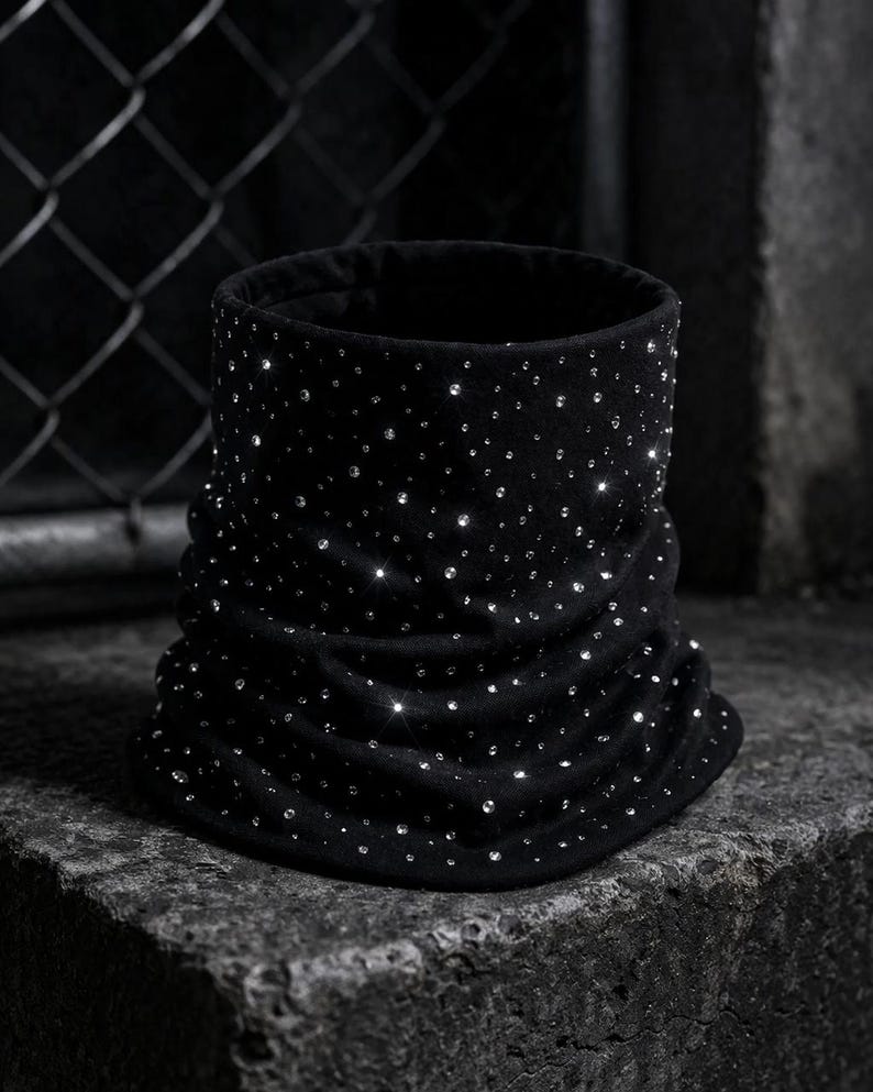 May include: A black, cylindrical neck warmer made of soft fabric, adorned with numerous sparkling rhinestones. The neck warmer has a ruched design and is displayed on a textured, dark surface.