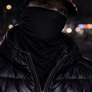 May include: A person wearing a black puffer jacket with a zipper, a black face covering, and a black baseball cap with a white stripe and a red brim. The background is dark with blurred lights.