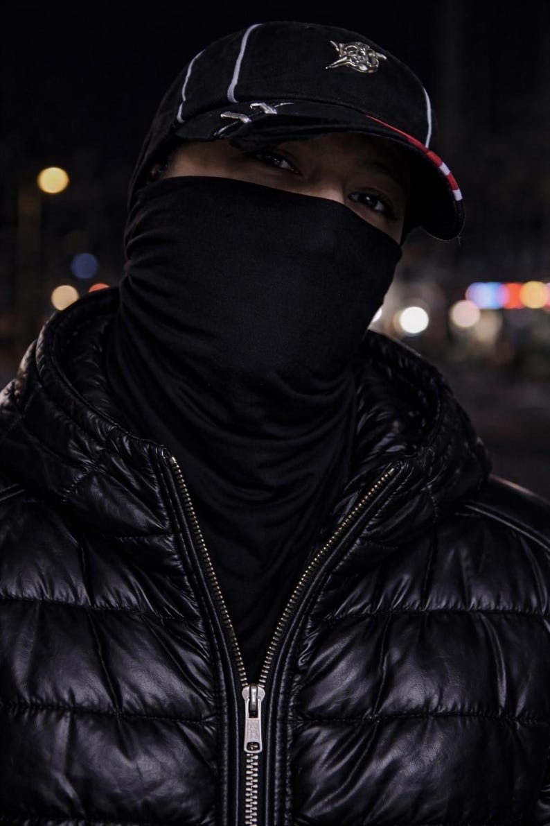 May include: A person wearing a black puffer jacket with a zipper, a black face covering, and a black baseball cap with a white stripe and a red brim. The background is dark with blurred lights.
