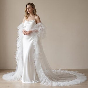 May include: A white satin slip dress with spaghetti straps and a cowl neckline. A sheer, ruffled, tulle wrap is draped over the shoulders and extends into a long train. The dress is floor-length and suitable for a wedding or formal event.