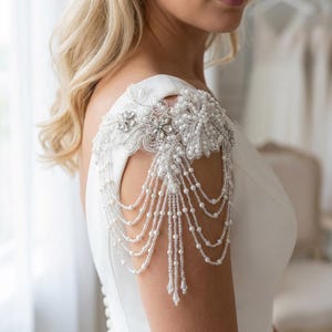 Könnte beinhalten: Ein weißes Brautschulter-Accessoire mit aufwendigen Perlen- und Perlendetails. Das Schulterstück hat ein florales Design mit herabhängenden Strängen aus Perlen und klaren Perlen, die dem Brautkleid einen Hauch von Eleganz verleihen.