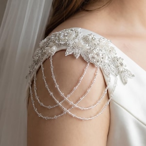 Könnte beinhalten: Ein weißes Brautschulter-Accessoire mit floralen und perlenbesetzten Verzierungen. Zarte Stränge aus klaren Perlen fallen den Arm hinunter. Das Schulterteil ist an einem weißen Kleid befestigt und sorgt für einen eleganten und romantischen Look.