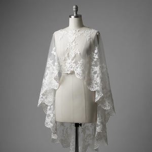 May include: A white lace bridal cape draped over a mannequin. The cape features intricate floral lace detailing around the neckline and edges, with a sheer, flowing design. The cape is a delicate, elegant accessory for weddings or special occasions.