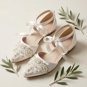 Puede incluir: Un par de bailarinas de satén marfil con puntera adornadas con perlas y pedrería. Las correas del tobillo se aseguran con lazos de satén. Hojas verdes dispersas alrededor de los zapatos, sugiriendo una boda u ocasión especial.