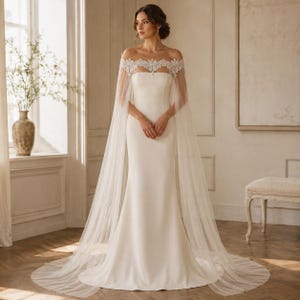 May include: A floor-length white gown featuring a strapless bodice and a sheer, floor-sweeping cape overlay. The cape is adorned with intricate white lace detailing across the shoulders and neckline, flowing into a long, translucent tulle train.