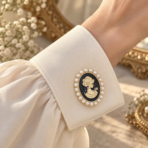 Puede incluir: Un puño de camisa de manga larga color crema con un broche ovalado decorativo. El broche tiene un fondo negro con una silueta de perfil en tono dorado de una persona, rodeado por un borde de pequeñas perlas blancas artificiales.