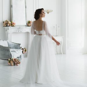 May include: A white bridal gown featuring a corset-style top with a lace-up back and a flowing tulle skirt. The dress has sheer sleeves and a long train. The setting is a bright, white room with a fireplace and a vintage-style chair.