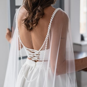 Puede incluir: Un vestido de novia blanco con espalda de corsé y falda fluida. El vestido presenta tirantes con cuentas de perlas y un velo transparente. La parte trasera del vestido es visible, mostrando el intrincado encaje del corsé. El estilo general es elegante y romántico.