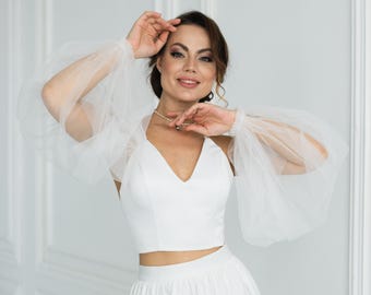 May include: A white two-piece bridal outfit featuring a crop top with a V-neck and a matching skirt. The outfit includes sheer, puffy sleeves. The model is wearing a pearl necklace.