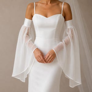 May include: A floor-length white gown featuring a sweetheart neckline, thin spaghetti straps, and detachable sheer, flowing bell sleeves that extend from the upper arms. The dress is constructed from a smooth, solid white fabric.