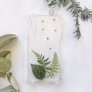K&ouml;nnte beinhalten: Eine transparente Handyh&uuml;lle mit einem Design aus gepressten Blumen. Die H&uuml;lle zeigt kleine wei&szlig;e Bl&uuml;ten und gr&uuml;ne Farne.