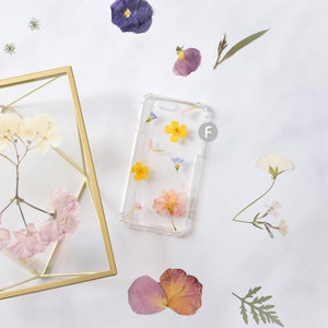 K&ouml;nnte beinhalten: Eine transparente Handyh&uuml;lle mit gepressten Blumen. Die H&uuml;lle zeigt gelbe, rosa und blaue Blumen. Die H&uuml;lle liegt auf einer wei&szlig;en Oberfl&auml;che mit anderen gepressten Blumen.
