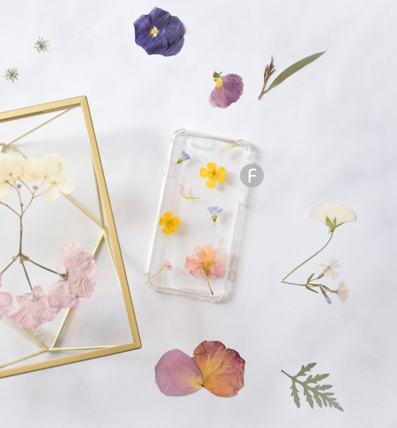 K&ouml;nnte beinhalten: Eine transparente Handyh&uuml;lle mit gepressten Blumen. Die H&uuml;lle zeigt gelbe, rosa und blaue Blumen. Die H&uuml;lle liegt auf einer wei&szlig;en Oberfl&auml;che mit anderen gepressten Blumen.