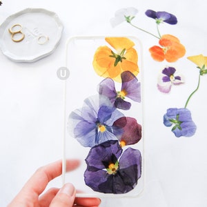 K&ouml;nnte beinhalten: Eine transparente Handyh&uuml;lle mit gepressten lila, gelben und orangefarbenen Blumen. Die Blumen sind vertikal in einer Reihe angeordnet.