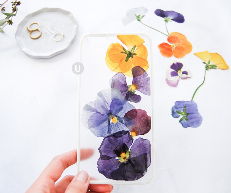 K&ouml;nnte beinhalten: Eine transparente Handyh&uuml;lle mit gepressten lila, gelben und orangefarbenen Blumen. Die Blumen sind vertikal in einer Reihe angeordnet.