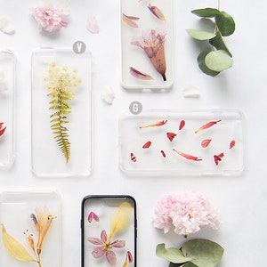 K&ouml;nnte beinhalten: Sechs transparente Handyh&uuml;llen mit gepressten Blumen und Bl&uuml;tenbl&auml;ttern darin. Die H&uuml;llen sind auf einer wei&szlig;en Oberfl&auml;che angeordnet. Die Blumen umfassen Rosa, Rot, Gelb und Wei&szlig;.