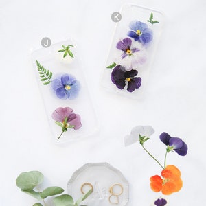 K&ouml;nnte beinhalten: Zwei transparente Handyh&uuml;llen mit gepressten Blumen. Die H&uuml;llen zeigen violette, blaue und wei&szlig;e Stiefm&uuml;tterchen. Eine kleine Schale mit zwei goldenen Ringen und dem Buchstaben "A" befindet sich in der unteren rechten Ecke.