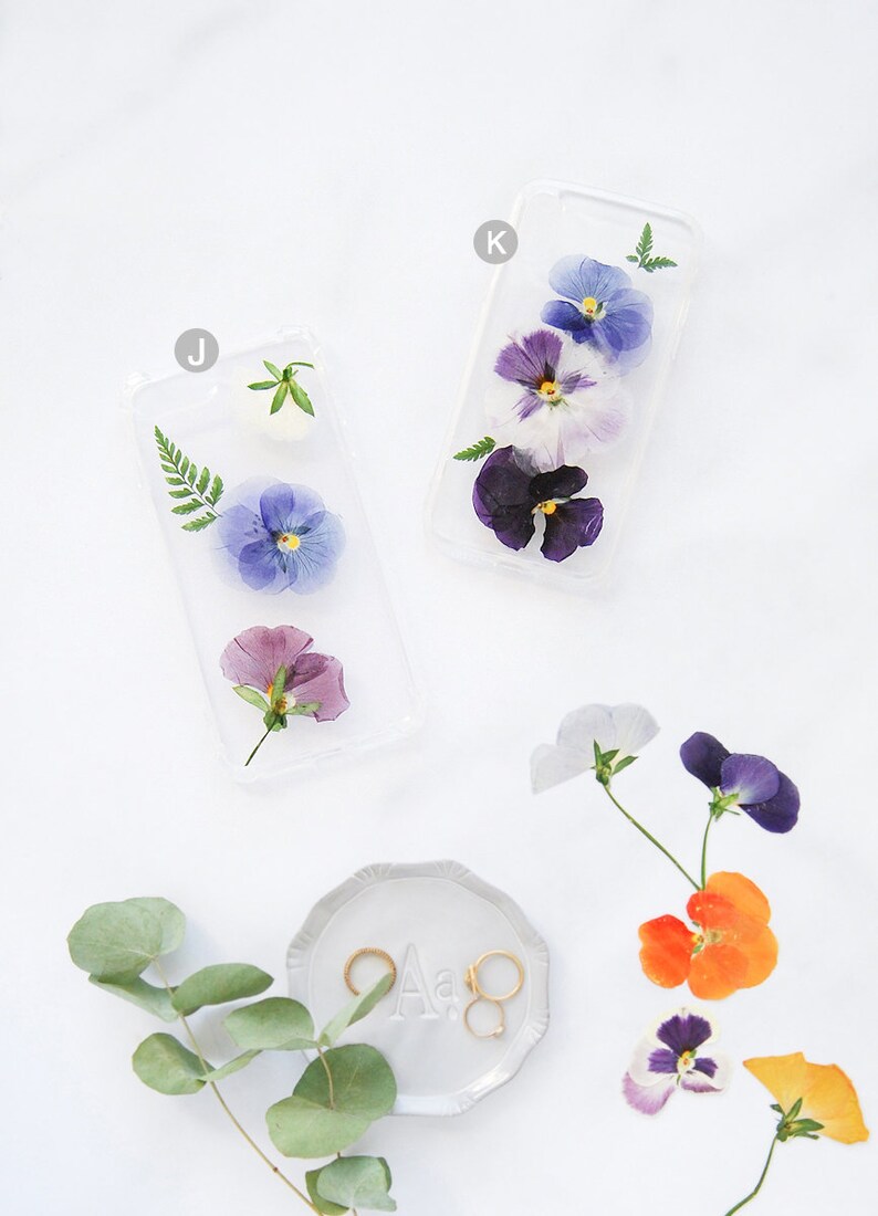 K&ouml;nnte beinhalten: Zwei transparente Handyh&uuml;llen mit gepressten Blumen. Die H&uuml;llen zeigen violette, blaue und wei&szlig;e Stiefm&uuml;tterchen. Eine kleine Schale mit zwei goldenen Ringen und dem Buchstaben "A" befindet sich in der unteren rechten Ecke.