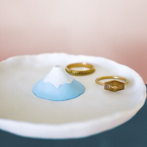 以下が含まれることがあります： 白磁製のリングディッシュ。青と白の山の形をしたリングホルダーと、金色のリングが2つあります。