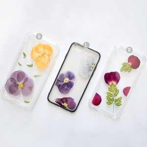 K&ouml;nnte beinhalten: Drei transparente Handyh&uuml;llen mit gepressten Blumen darin. Die H&uuml;llen sind transparent und zeigen die Blumen im Detail. Die Blumen sind lila, gelb und rot.