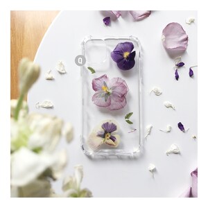 K&ouml;nnte beinhalten: Eine transparente Handyh&uuml;lle mit drei gepressten lila und wei&szlig;en Blumen darin. Die Blumen sind vertikal in der H&uuml;lle angeordnet.