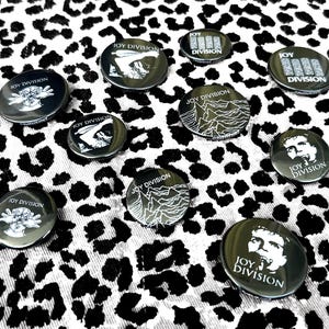 Puede incluir: Una colección de botones de la banda Joy Division en blanco y negro. Los botones presentan varios gráficos y el nombre de la banda. Están dispuestos sobre una tela con estampado de leopardo en blanco y negro.