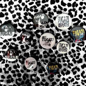 Op de afbeelding: Een verzameling van tien ronde speldjes met diverse Fugazi-bandafbeeldingen en tekst. De ontwerpen bevatten zwart-wit portretten van de band, het woord Fugazi in vetgedrukte letters en de albumtitel Repeater in zwarte tekst op een witte achtergrond.