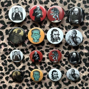 Peut inclure: Une collection de quatorze badges ronds à épingle présentant des illustrations de monstres classiques de films d'horreur. Les motifs incluent des représentations du monstre de Frankenstein, de Dracula, du loup-garou et de la momie.