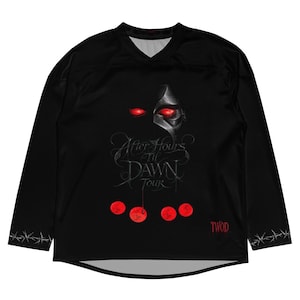 May include: A black long-sleeve jersey with a V-neckline. The front features a graphic of a metallic mask with glowing red eyes, the text "After Hours til DAWN Tour," four red circular icons, and the red text "TWOD." Barbed wire patterns line the cuffs.