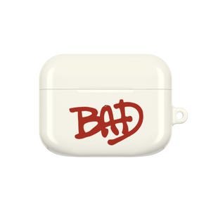 May include: A cream-colored protective case for wireless earbuds featuring a smooth, rounded rectangular shape. The front of the case displays the word BAD printed in a bold, dark red, hand-drawn graffiti-style font with a small circular cutout.