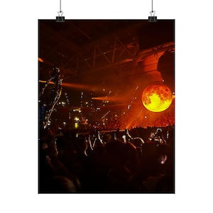 May include: A concert poster featuring a large, illuminated sphere resembling a fiery moon suspended above a crowd. The scene is bathed in warm orange and yellow light, with silhouettes of people in the foreground.