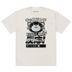 May include: Cream-colored t-shirt with a graphic design featuring Japanese text and a cartoon bear face. The design includes the words "KISS LAND" and "反応!" in bold, black lettering. The shirt has a crew neck and short sleeves.