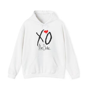 May include: White hoodie with a front pocket and a drawstring hood. The front features a black "XO" graphic with a red heart and the text "The Weeknd" in black cursive.