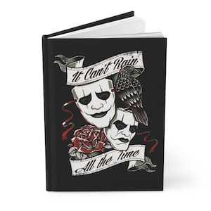 May include: A black hardcover journal featuring a graphic illustration of two white theatrical masks, a black crow, and a red rose. The design includes two banners with the text It Can't Rain All the Time in a black script font against the dark background.